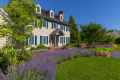 This charming two-story house, with beige siding and blue shutters, is nestled in lush greenery. Vibrant flowers and expertly designed hardscaping services in Delaware County, PA enhance its traditional appeal, complete with brick pathways under a clear blue sky.
