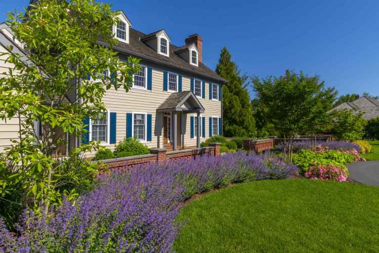 This charming two-story house, with beige siding and blue shutters, is nestled in lush greenery. Vibrant flowers and expertly designed hardscaping services in Delaware County, PA enhance its traditional appeal, complete with brick pathways under a clear blue sky.