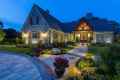 A beautifully illuminated stone house at dusk, featuring gabled roofs and wooden accents. This Delaware County, PA gem showcases expertly crafted hardscaping services with a curved stone walkway, flowering plants, and large pots nestled in a lush lawn amid a tranquil natural setting.