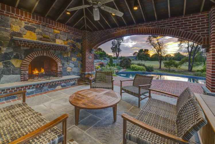 Experience the ultimate in outdoor living in Delaware County, PA, with a stone fireplace illuminating wicker furniture. This charming patio overlooks a pool and open field with sunset views through an arched brick opening, enhanced by a ceiling fan and wooden beams.