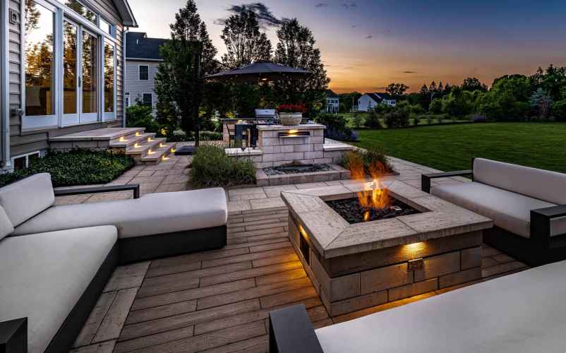 Outdoor living at its finest in Delaware County, PA, with cushioned seating around a lit fire pit. Steps with lighting guide you to the house, and a barbecue station with an umbrella is nearby. Overlooking a large lawn and garden beneath a sunset's warm glow, it's the perfect retreat.