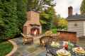 A patio featuring an inviting lit outdoor fireplace is surrounded by wicker furniture. A round metal table is elegantly set for four with plates, wine glasses, and a bottle. Lush greenery and trees from top-notch landscaping services in Delaware County PA create a serene backdrop.