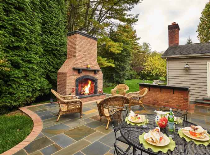 A patio featuring an inviting lit outdoor fireplace is surrounded by wicker furniture. A round metal table is elegantly set for four with plates, wine glasses, and a bottle. Lush greenery and trees from top-notch landscaping services in Delaware County PA create a serene backdrop.