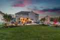 Backyard view of a large house at sunset in Delaware County, PA. The patio is illuminated, featuring outdoor seating, a fire pit, and a stone fireplace. There's expertly maintained landscaping with colorful plants and a dramatic sky with pink and blue hues overhead.