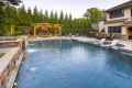 A luxurious backyard in Delaware County, PA, showcases a large swimming pool with water jets and stone decking. A wooden pergola with a fireplace and seating area accentuates the space, while lounge chairs partially submerged in the pool invite relaxation against a backdrop of expertly crafted hardscaping.