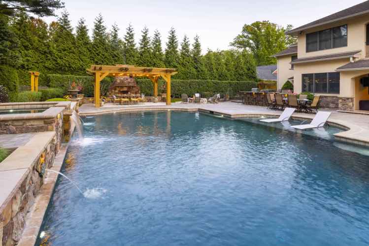 A luxurious backyard in Delaware County, PA, showcases a large swimming pool with water jets and stone decking. A wooden pergola with a fireplace and seating area accentuates the space, while lounge chairs partially submerged in the pool invite relaxation against a backdrop of expertly crafted hardscaping.