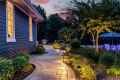 A beautifully landscaped path, crafted through expert landscaping services in Delaware County PA, curves alongside a house at dusk, illuminated by warm lighting. Lined with green shrubs, it leads to a pool area with a blue umbrella. Trees and a vibrant sunset complete this serene scene.