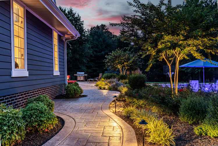 A beautifully landscaped path, crafted through expert landscaping services in Delaware County PA, curves alongside a house at dusk, illuminated by warm lighting. Lined with green shrubs, it leads to a pool area with a blue umbrella. Trees and a vibrant sunset complete this serene scene.