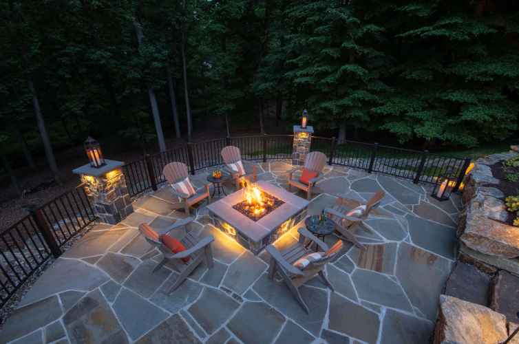 An outdoor patio in Delaware County, PA, surrounded by a metal railing, features a lit firepit and Adirondack chairs with cushions. Lanterns and stone flooring enhance the cozy ambiance, perfectly blending nature with expertly crafted hardscaping services for inviting outdoor living.