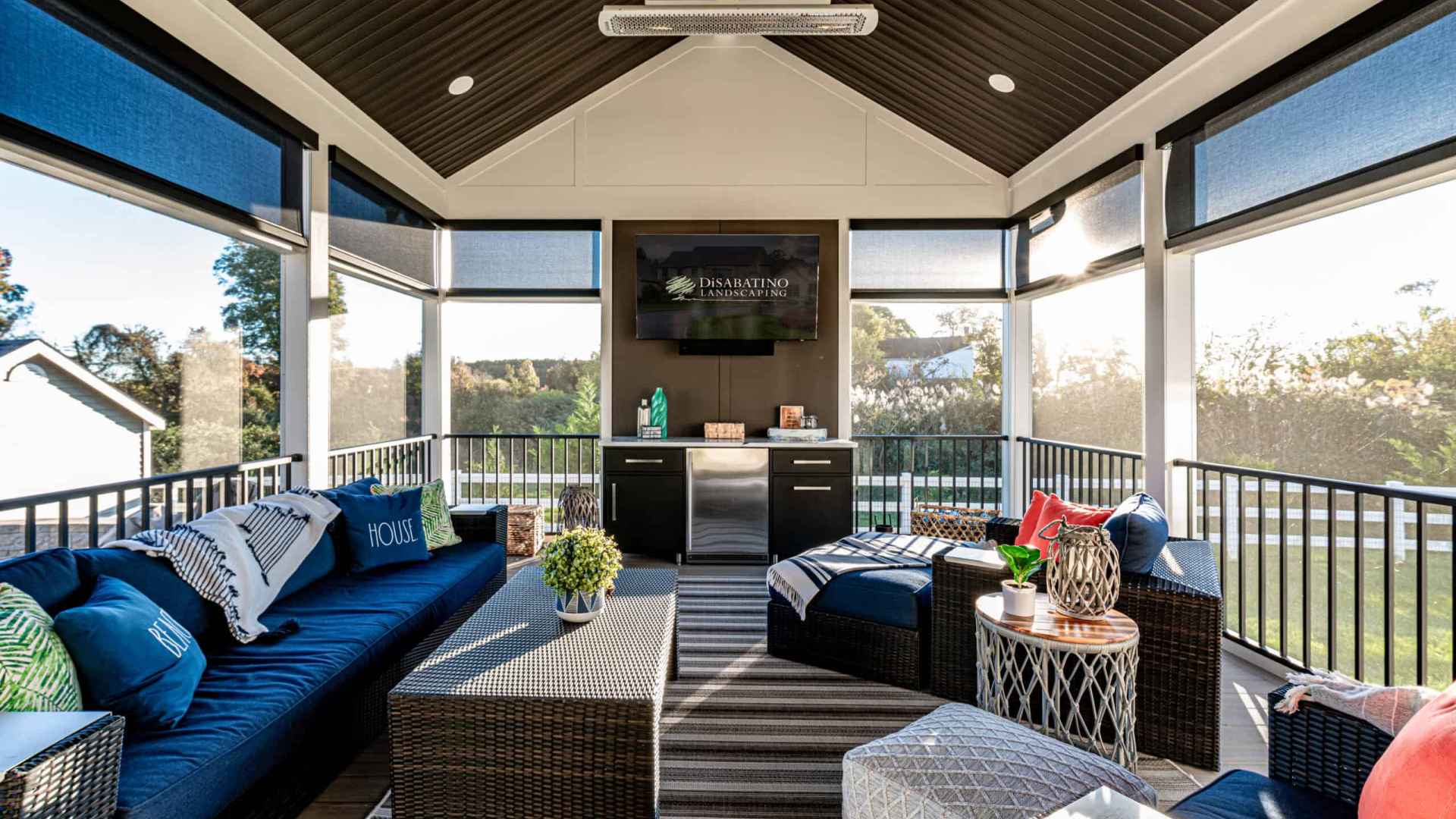 Stylish sunroom with dark wicker furniture, blue cushions, and a striped rug sets the stage for outdoor living in Delaware County, PA. Large windows offer scenic views and ample natural light. A small table with decorative items and plants completes the cozy space.