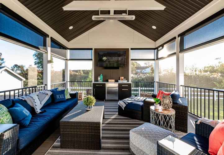Stylish sunroom with dark wicker furniture, blue cushions, and a striped rug sets the stage for outdoor living in Delaware County, PA. Large windows offer scenic views and ample natural light. A small table with decorative items and plants completes the cozy space.