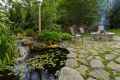 A serene backyard scene in Delaware County, PA, with a stone patio showcases the beauty of outdoor living. It features a smokey fire pit, two chairs, a lush garden, and a pond with lily pads and a small waterfall. Surrounded by trees, this tranquil spot offers an inviting retreat.