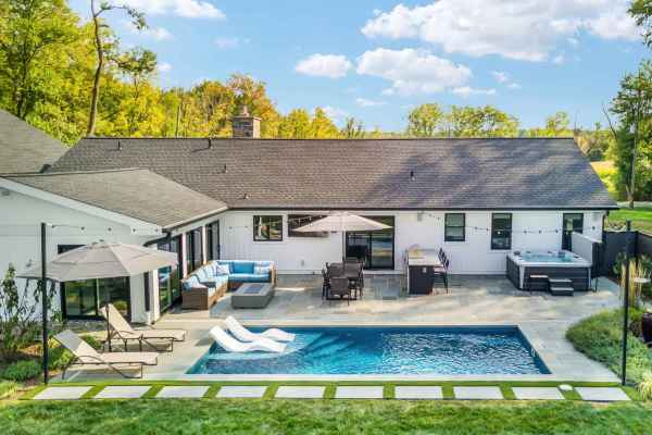 Aerial view of a modern backyard in Delaware County, PA, featuring stunning hardscaping services. A swimming pool is surrounded by a stone patio with lounge chairs, a dining set under an umbrella, a hot tub, and sectional sofa. The white house has a gray roof with trees in the background.
