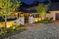 A warmly lit stone house with glowing lanterns graces the gate pillars. The brick pathway, crafted with expert hardscaping services in Delaware County PA, is lined with small trees and flower beds leading to the entrance. Cobblestone pavement frames this inviting outdoor living space under a dark evening sky.