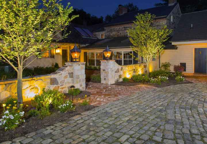 A warmly lit stone house with glowing lanterns graces the gate pillars. The brick pathway, crafted with expert hardscaping services in Delaware County PA, is lined with small trees and flower beds leading to the entrance. Cobblestone pavement frames this inviting outdoor living space under a dark evening sky.