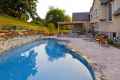 A luxurious backyard features a curved pool with a stone border, enhanced by expert landscaping services in Delaware County, PA. Surrounded by stylish patio furniture and a pergola with outdoor seating, the serene setting includes a stone wall lining the manicured garden.