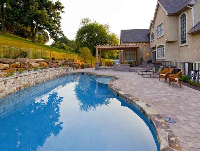 A luxurious backyard features a curved pool with a stone border, enhanced by expert landscaping services in Delaware County, PA. Surrounded by stylish patio furniture and a pergola with outdoor seating, the serene setting includes a stone wall lining the manicured garden.