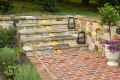 Stone steps with lanterns lead up from a brick patio, enveloped by a stone wall. Small plants and flowers grace the foreground, while soft yellow lights illuminate the steps. Lush greenery is visible in the background, creating an enchanting outdoor living space in Delaware County, PA.