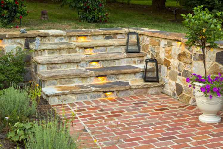 Stone steps with lanterns lead up from a brick patio, enveloped by a stone wall. Small plants and flowers grace the foreground, while soft yellow lights illuminate the steps. Lush greenery is visible in the background, creating an enchanting outdoor living space in Delaware County, PA.
