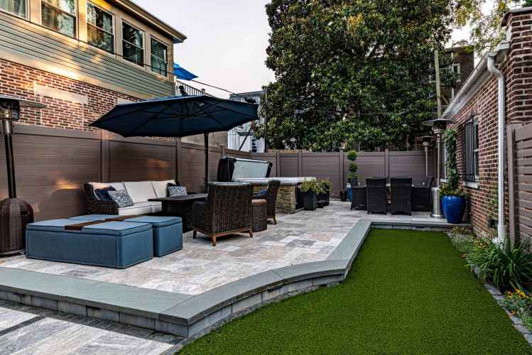 A spacious patio highlights outdoor living in Delaware County, PA, featuring wicker chairs and a large umbrella. A dining table sits nearby. The area is embraced by wooden fencing and a lush artificial grass lawn, framed by brick walls.