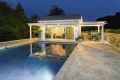 Elegant modern pool house illuminated at night with a pergola and glass doors. Surrounded by a beautifully designed patio and adjacent to a swimming pool, it reflects the structure. Outdoor furniture enhances the outdoor living vibe of Delaware County, PA, with trees visible in the background.