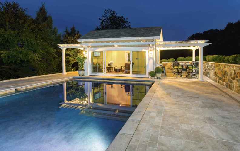 Elegant modern pool house illuminated at night with a pergola and glass doors. Surrounded by a beautifully designed patio and adjacent to a swimming pool, it reflects the structure. Outdoor furniture enhances the outdoor living vibe of Delaware County, PA, with trees visible in the background.
