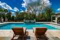 Two wooden lounge chairs face a pristine, rectangular swimming pool under a partly cloudy blue sky. The pool is surrounded by a stone patio and lush greenery, showcasing the finest hardscaping services Delaware County PA has to offer for an idyllic outdoor living experience.