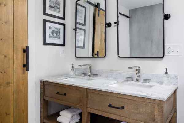 Modern bathroom with a wooden double vanity featuring a marble countertop and two black-framed mirrors. Towels rest on open shelves below. A sliding barn door and framed black and white photos adorn the walls, while the pebble-like floor pattern infuses an outdoor living aesthetic popular in Delaware County, PA.