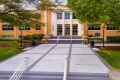 A brick academic building with large windows and white columns is surrounded by green trees, enhanced by expert landscaping services in Delaware County, PA. A wide stone walkway with steps leads to the entrance, featuring lamps on either side. The scene is bathed in soft daylight.