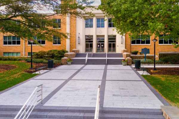 A brick academic building with large windows and white columns is surrounded by green trees, enhanced by expert landscaping services in Delaware County, PA. A wide stone walkway with steps leads to the entrance, featuring lamps on either side. The scene is bathed in soft daylight.