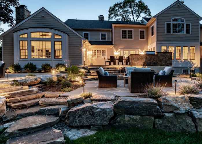 A spacious backyard at dusk features a stylish patio with wicker furniture. The house's large windows and warm lighting illuminate the area, while stone steps and boulders accentuate the landscaped garden. Perfect for outdoor living in Delaware County, PA.