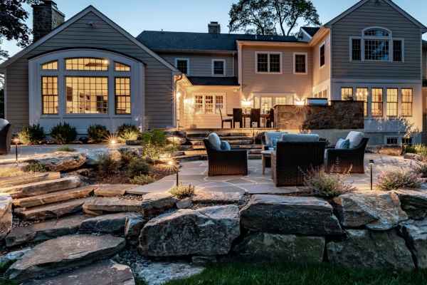 A spacious backyard at dusk features a stylish patio with wicker furniture. The house's large windows and warm lighting illuminate the area, while stone steps and boulders accentuate the landscaped garden. Perfect for outdoor living in Delaware County, PA.