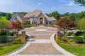 A large, elegant house with a stone facade stands at the end of a circular driveway. Surrounded by expertly crafted gardens and neatly trimmed hedges, thanks to premier landscaping services Delaware County PA, it basks under the blue sky and lush greenery in this serene setting.