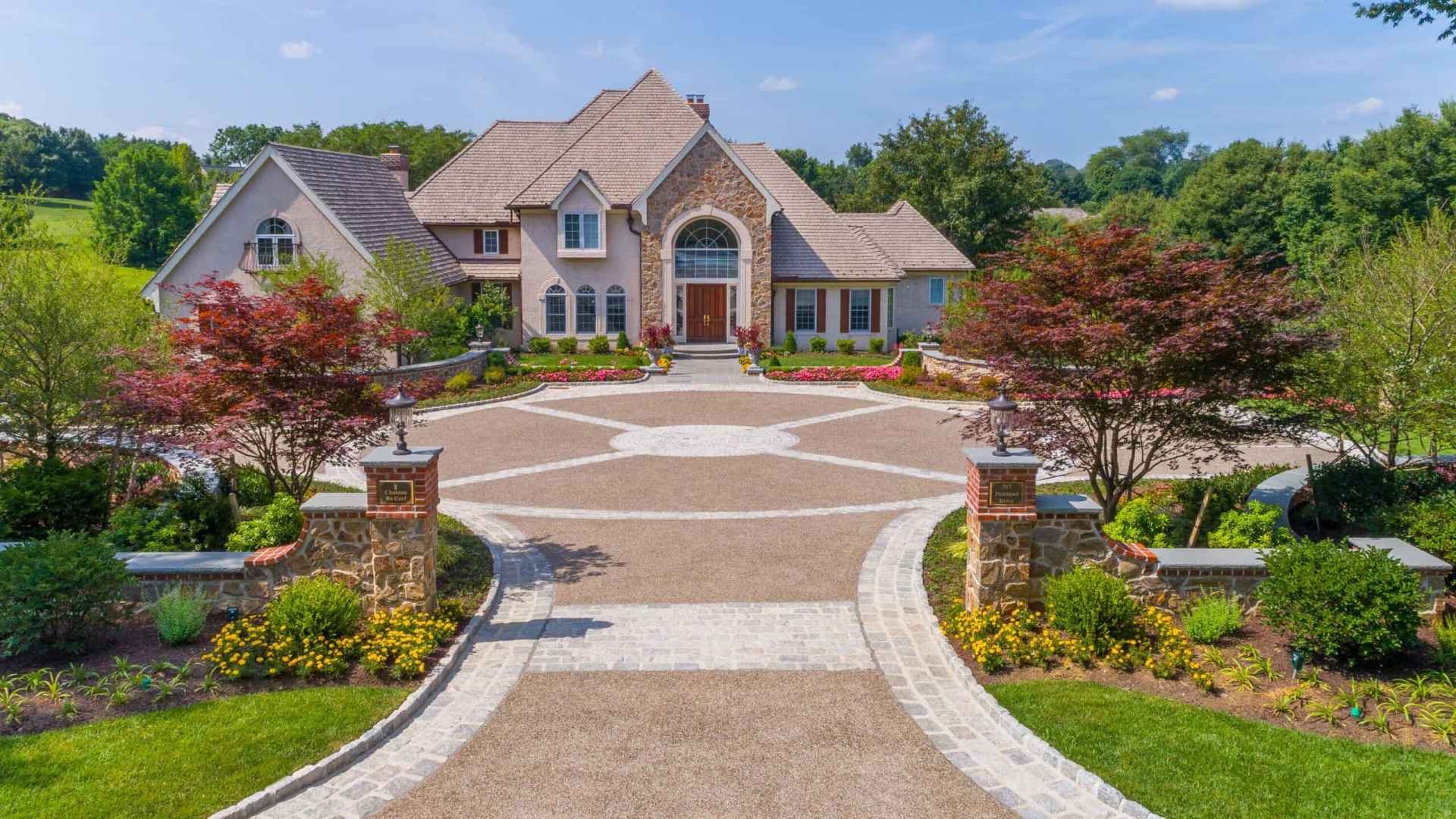 A large, elegant house with a stone facade stands at the end of a circular driveway. Surrounded by expertly crafted gardens and neatly trimmed hedges, thanks to premier landscaping services Delaware County PA, it basks under the blue sky and lush greenery in this serene setting.