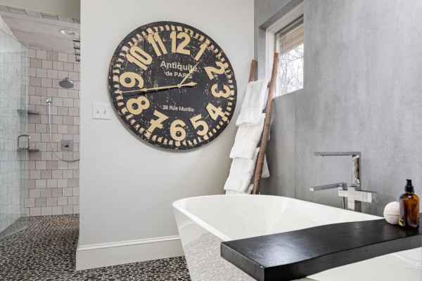 Modern bathroom with a freestanding white tub and large black and white wall clock, reminiscent of the elegance found in top landscaping services Delaware County PA offers. Pebble-style tiles cover the floor, complemented by a towel ladder rack. The shower area boasts white tiled walls and a sleek glass door.