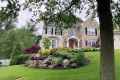 A two-story house with stone and beige siding stands amidst lush landscaping, a testament to premier landscaping services in Delaware County, PA. The front yard boasts a manicured lawn, vibrant flower beds, rocks, and trees under a cloudy sky, with a white fence on the left.