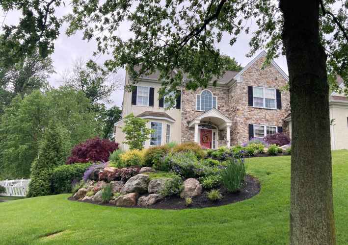 A two-story house with stone and beige siding stands amidst lush landscaping, a testament to premier landscaping services in Delaware County, PA. The front yard boasts a manicured lawn, vibrant flower beds, rocks, and trees under a cloudy sky, with a white fence on the left.