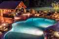 In a serene evening ambiance, an illuminated backyard scene showcases a glowing pool with a waterfall. Beside it, a rustic pavilion featuring a bar and string lights enhances outdoor living in Delaware County, PA. Landscaped gardens and trees surround the area, completing the picturesque setting.