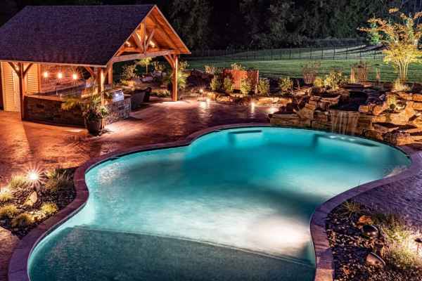 In a serene evening ambiance, an illuminated backyard scene showcases a glowing pool with a waterfall. Beside it, a rustic pavilion featuring a bar and string lights enhances outdoor living in Delaware County, PA. Landscaped gardens and trees surround the area, completing the picturesque setting.