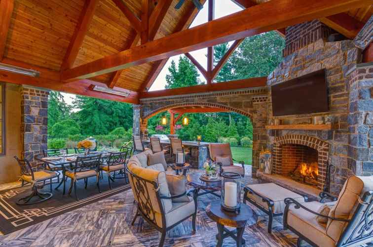 Luxurious outdoor patio with wooden ceiling and stone walls, featuring cushioned seating around a fireplace with a TV above. This exquisite outdoor living space in Delaware County, PA, includes a large dining table beside lush garden views, creating an inviting atmosphere with warm lighting.