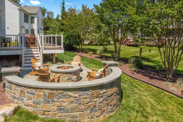 Adirondack chairs encircle a stone fire pit on a patio, enhancing the outdoor living experience in this Delaware County, PA backyard. Bordered by lush grass and pristine landscaping, the scene features a white house with a deck under a clear blue sky.