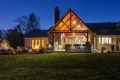 A large stone house with a wooden patio is warmly lit against a twilight sky in Delaware County, PA. The patio features a fireplace and seating area, perfect for outdoor living. The surrounding yard boasts expert landscaping with trees and shrubs, enhancing its natural beauty.