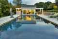 The rectangular pool with clear water reflects a yellow pool house and white pergola. A sculpture is on the left, while trees, bushes, and expertly designed hardscaping services in Delaware County, PA, create a serene escape with lounge chairs on the stone patio to the right.