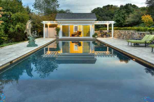 The rectangular pool with clear water reflects a yellow pool house and white pergola. A sculpture is on the left, while trees, bushes, and expertly designed hardscaping services in Delaware County, PA, create a serene escape with lounge chairs on the stone patio to the right.