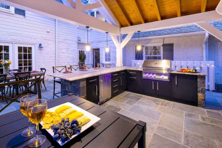 Outdoor living in Delaware County, PA, comes to life with this exquisite setup: a kitchen boasting modern appliances, slate flooring, and wooden ceiling. A bar counter with crackers, cheese, grapes, and wine awaits. A patio dining area is visible amidst refined landscaping services.