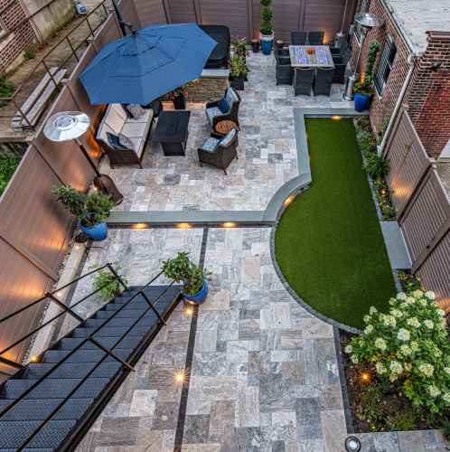 Aerial view of a modern patio in Delaware County, PA, with stone tiles, artificial grass, and cozy seating. Highlighted by a black metal staircase, blue planters, decorative lighting, and a dining set under a dark umbrella. Brick walls wrap around this charming outdoor living space.