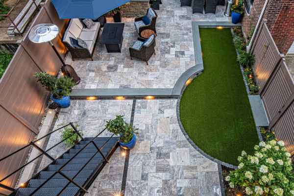 Aerial view of a modern patio in Delaware County, PA, with stone tiles, artificial grass, and cozy seating. Highlighted by a black metal staircase, blue planters, decorative lighting, and a dining set under a dark umbrella. Brick walls wrap around this charming outdoor living space.