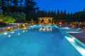 The illuminated backyard pool at dusk, featuring a waterfall and stone patio, highlights the exquisite outdoor living in Delaware County PA. Lounge chairs sit to the right, while a lit gazebo offers seating in the background, bordered by tall trees.