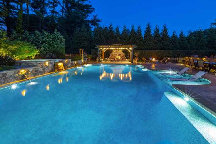 The illuminated backyard pool at dusk, featuring a waterfall and stone patio, highlights the exquisite outdoor living in Delaware County PA. Lounge chairs sit to the right, while a lit gazebo offers seating in the background, bordered by tall trees.