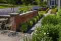 A garden path with lush greenery and blooming white daisies leads to a brick wall. Lavender plants and trimmed bushes line the walkway. An outdoor seating area with metal chairs sits near the path, under a clear blue sky, showcasing expert landscaping services in Delaware County PA.